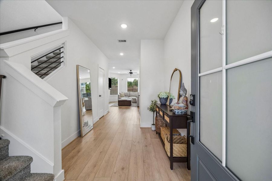 A welcoming foyer features soaring ceilings, durable Shaw vinyl plank flooring, and a direct sightline to the wall of windows overlooking the backyard. A conveniently tucked-away powder room beneath the stairs adds function and style.