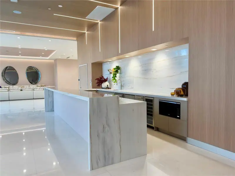 Custom wet bar featuring a stone waterfall-edge island, stone slab backsplash, light wood-finish cabinetry, integrated linear ceiling lighting, and built-in refrigeration