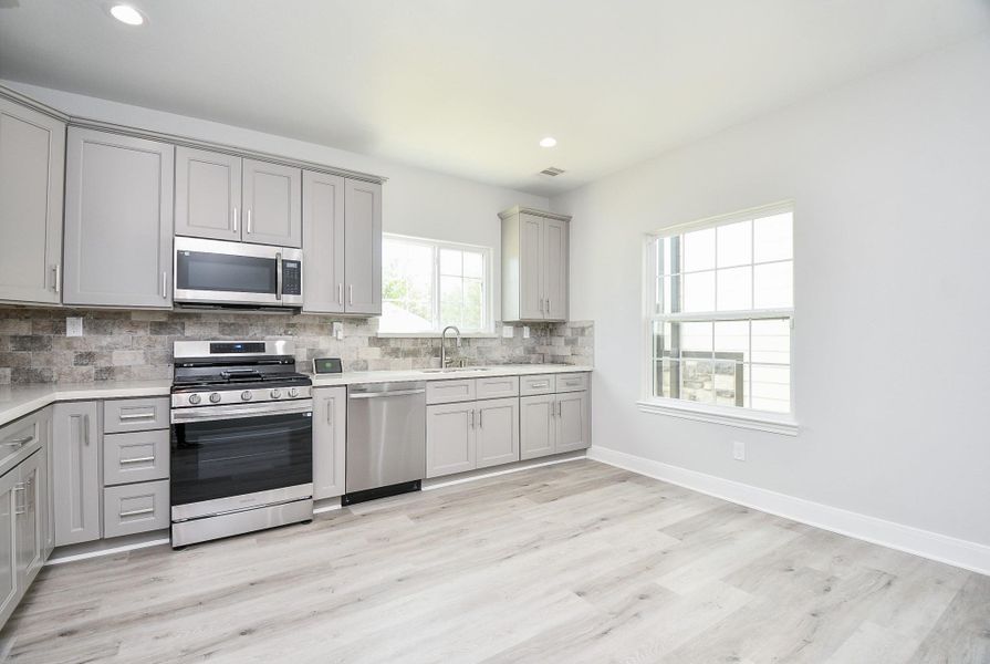 This kitchen features modern gray cabinetry, stainless steel appliances, and a sleek tile backsplash. The space is bright with ample natural light from large windows, complemented by light wood flooring for an airy feel.