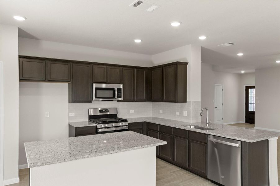 Kitchen featuring appliances with stainless steel finishes, a peninsula, decorative backsplash, dark brown cabinetry, and light stone countertops