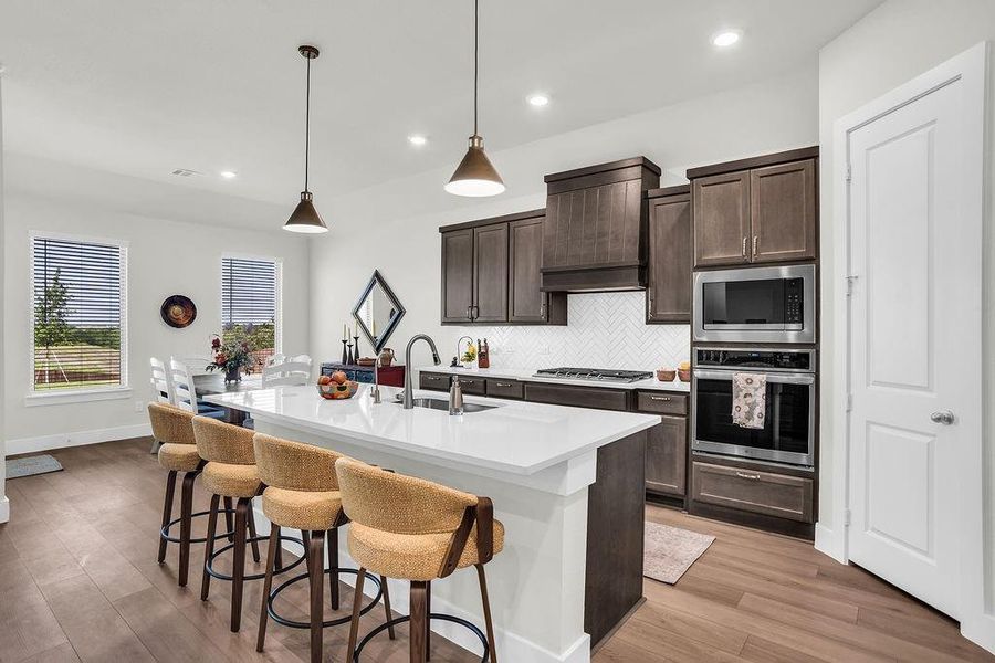 Kitchen featuring a substantial island with breakfast bar, undermount sink with gooseneck brushed nickel faucet, wood-finish flooring, dark wood cabinetry, a white herringbone tile backsplash, walk-in pantry and stainless steel appliances including a propane gas cooktop, electric built-in oven and microwave open to living and breakfast rooms