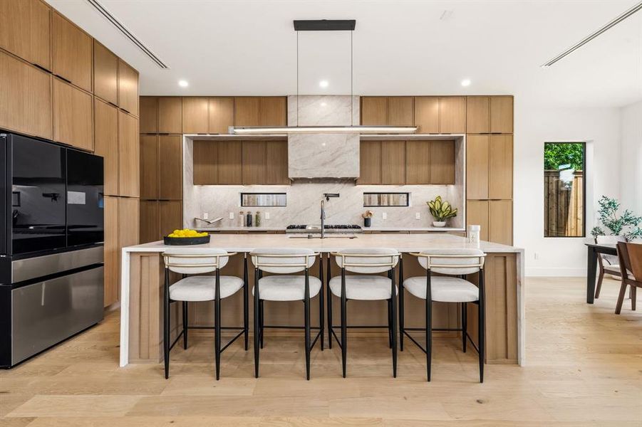Gourmet kitchen featuring a large central island with seating, integrated appliances, wood-finish cabinetry, a striking marble backsplash, and under-cabinet lighting