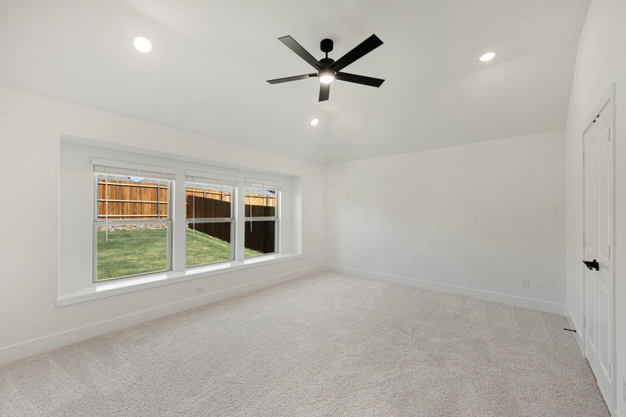 Empty bedroom with carpet flooring, black ceiling fan, and triple windows overlooking a backyard
