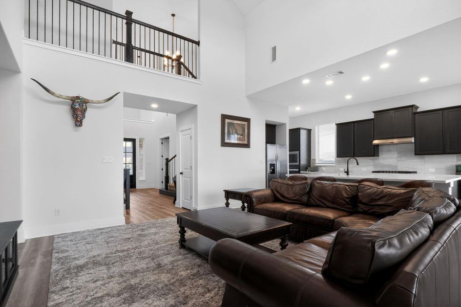 Open-concept living area featuring high ceilings, wood-finish flooring, a second-story interior balcony with black railing, recessed lighting, and a kitchen with dark cabinetry and a light-toned countertop Open-concept living area featuring high ceilings, wood-finish flooring, a second-story interior balcony with black railing, recessed lighting, and a kitchen with dark cabinetry and a light-toned countertop
