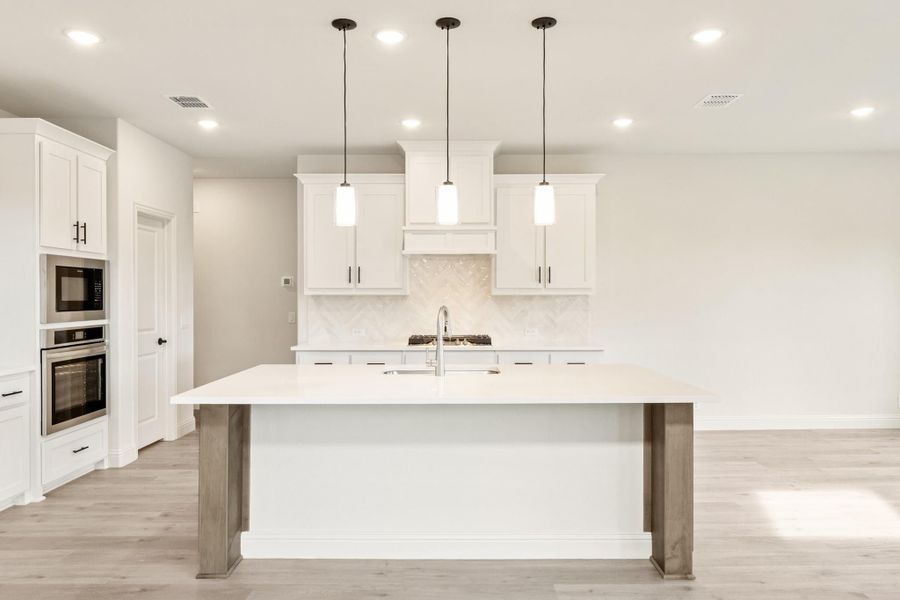 White kitchen with large center island, pendant lights, white cabinets, and stainless steel appliances on light hardwood floors White kitchen with large center island, pendant lights, white cabinets, and stainless steel appliances on light hardwood floors