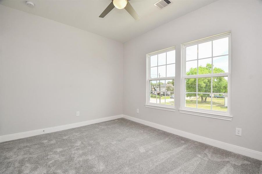 This is a bright, spacious bedroom featuring plush carpeting, neutral walls, a ceiling fan, and large windows offering plenty of natural light and a view of the outside greenery.