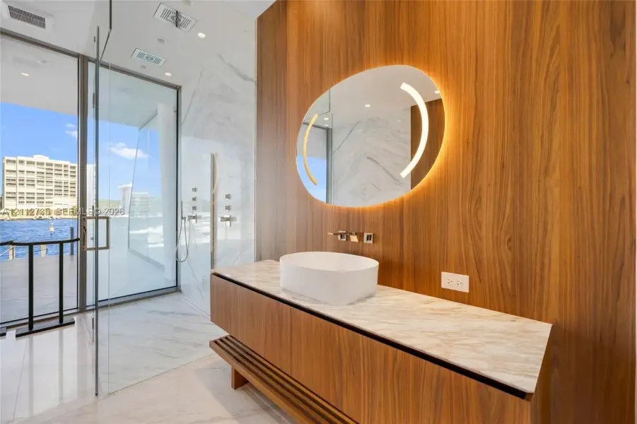 Floating wood-finish vanity with a stone-top surface and vessel sink, illuminated oval mirror, floor-to-ceiling glass-enclosed shower, and polished stone-finish flooring