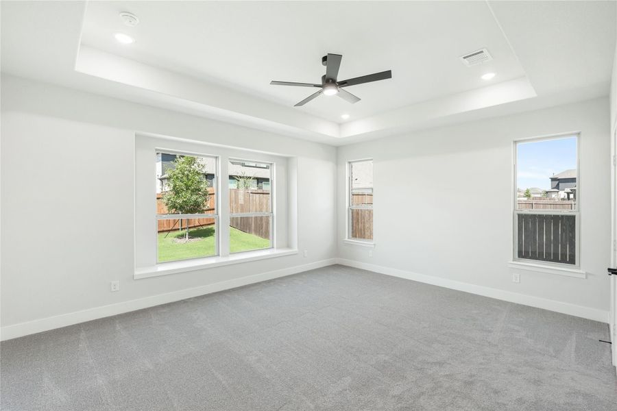 Empty bedroom with tray ceiling, ceiling fan, gray carpet, and large windows overlooking backyard Empty bedroom with tray ceiling, ceiling fan, gray carpet, and large windows overlooking backyard