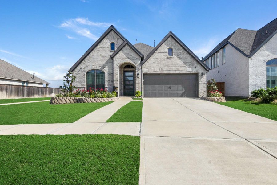 This photo showcases a modern, single-story brick home with a gabled roof and a two-car garage. The front yard is well-maintained, featuring lush green grass and neatly arranged flower beds. A wide driveway and a clear blue sky complete the inviting exterior.