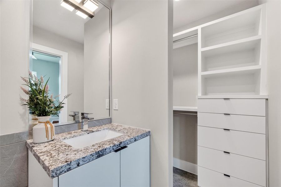 The first-floor bath vanity features a stone-look countertop with undermount sink and modern faucet. An adjacent walk-in closet, visible in the background, is outfitted with built-in shelving and a drawer unit, providing ample storage for a first-floor bedroom suite.