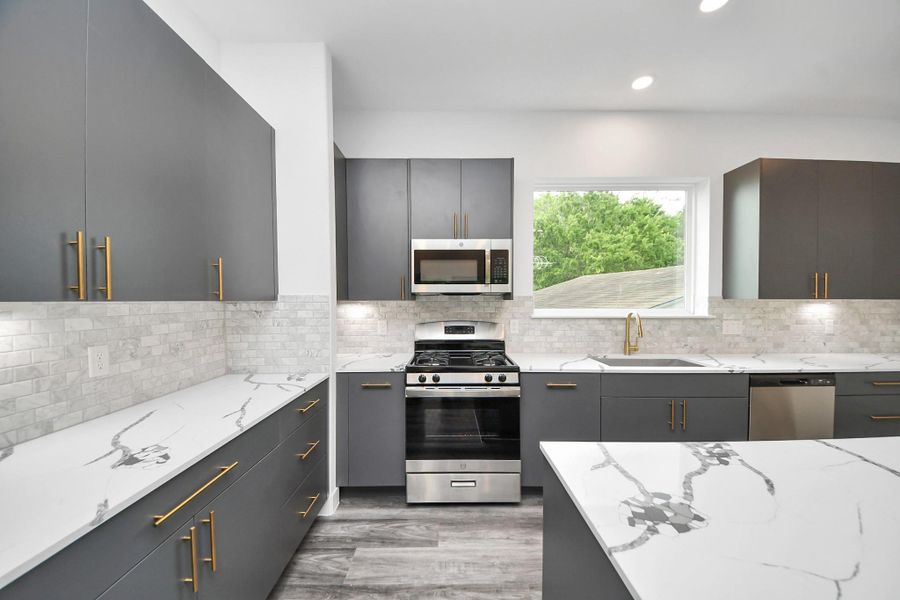 Modern kitchen with sleek gray cabinets, gold hardware, and marble-style countertops. Features include stainless steel appliances, a gas stove, and a large window offering natural light. Modern kitchen with sleek gray cabinets, gold hardware, and marble-style countertops. Features include stainless steel appliances, a gas stove, and a large window offering natural light.