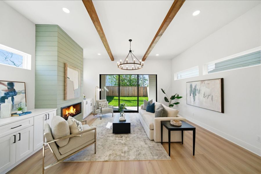 This living room features a modern design with high ceilings and exposed wooden accent beams. It includes a cozy fireplace, large windows for natural light, and sliding glass doors that open to a lush backyard. The space is elegantly furnished, offering a warm and inviting atmosphere.