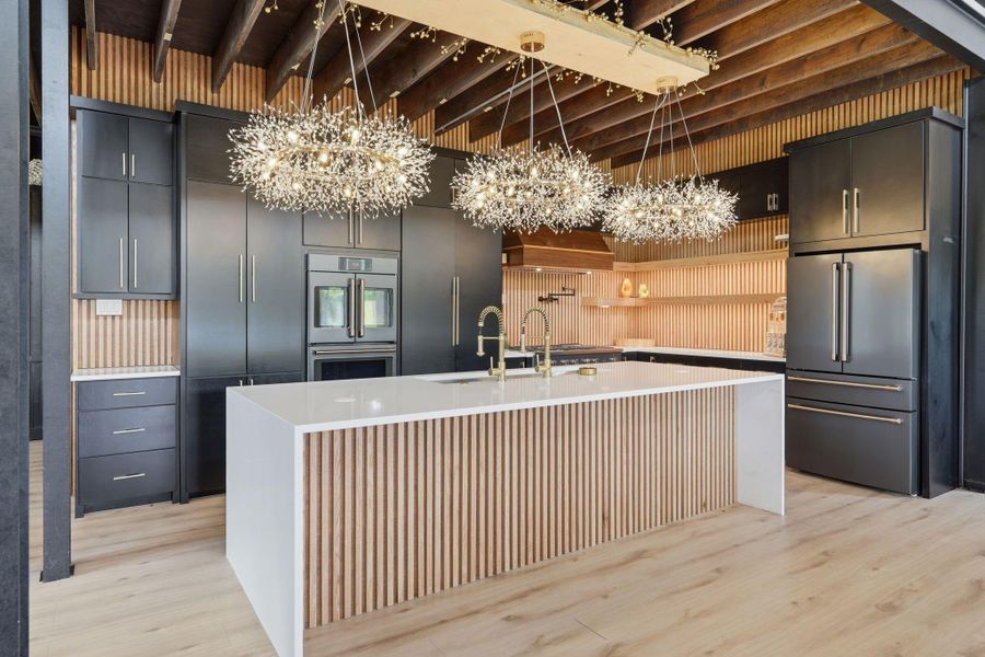 This modern kitchen features sleek black cabinetry, a spacious white island with a stylish wood panel design, and luxurious lighting fixtures. It includes high-end stainless steel appliances and warm wood accents, creating an inviting, contemporary atmosphere.