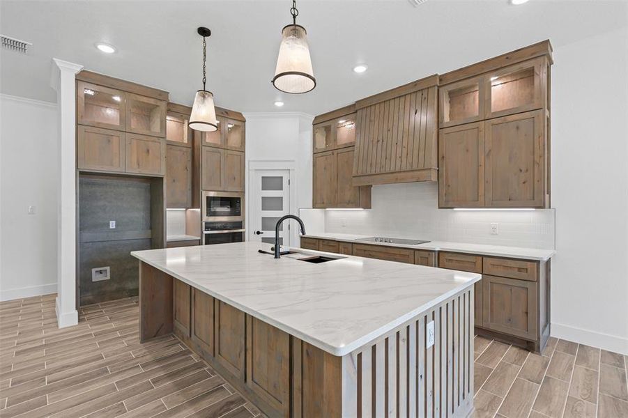 Kitchen with ornamental molding, light wood-style floors, a center island with sink, stainless steel oven, and decorative light fixtures Kitchen with ornamental molding, light wood-style floors, a center island with sink, stainless steel oven, and decorative light fixtures
