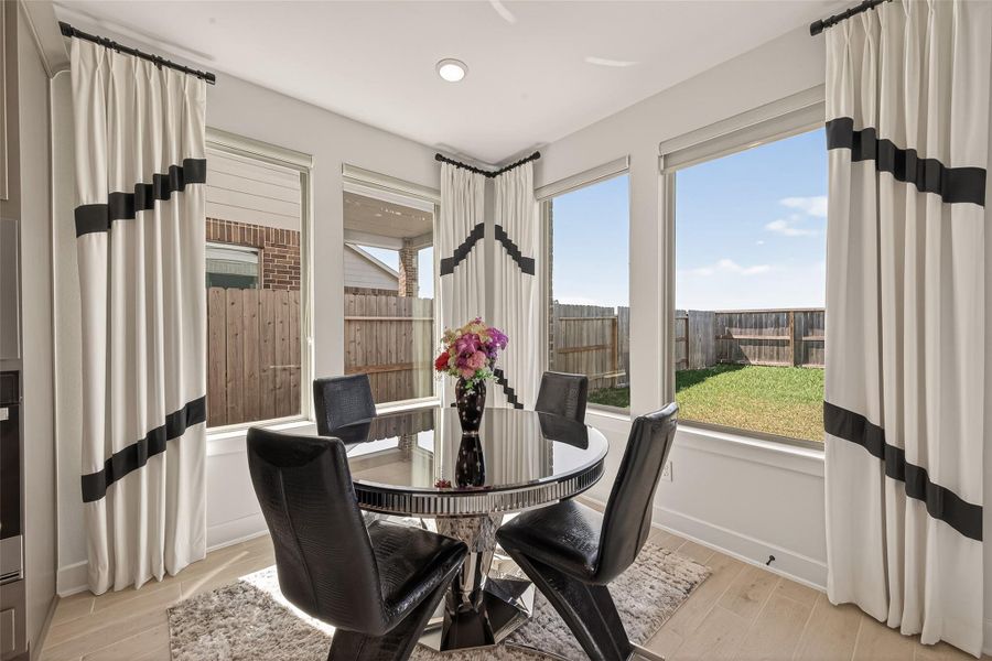 This bright dining area features large windows with elegant curtains, a modern round glass table, and sleek black chairs, offering a cozy space with views of the fenced backyard.