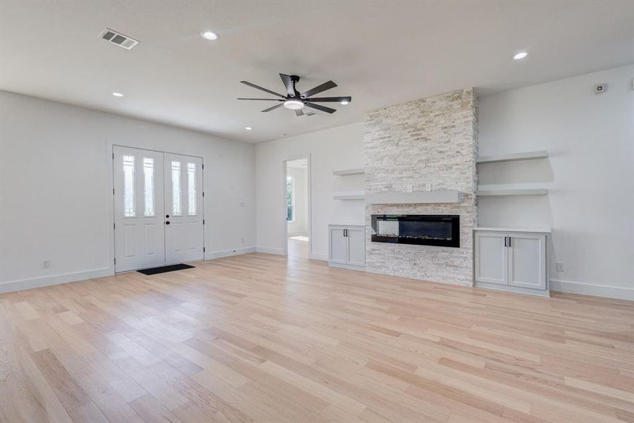 Spacious living area featuring light wood-finish flooring, a stone-clad fireplace with an electric insert, built-in cabinetry, floating shelves, and recessed lighting Spacious living area featuring light wood-finish flooring, a stone-clad fireplace with an electric insert, built-in cabinetry, floating shelves, and recessed lighting