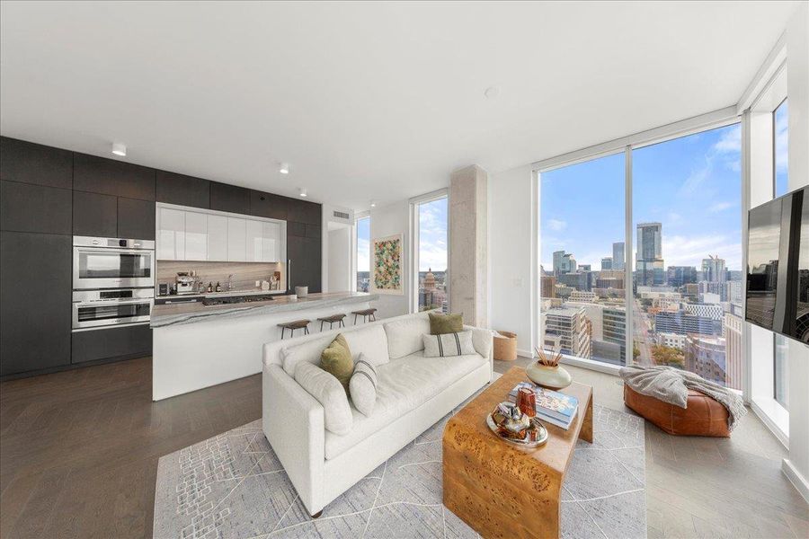 Light-filled living area with panoramic skyline views, opening to the chef’s kitchen featuring upgraded Fantasy Ocean marble countertops and sleek custom cabinetry. Light-filled living area with panoramic skyline views, opening to the chef’s kitchen featuring upgraded Fantasy Ocean marble countertops and sleek custom cabinetry.