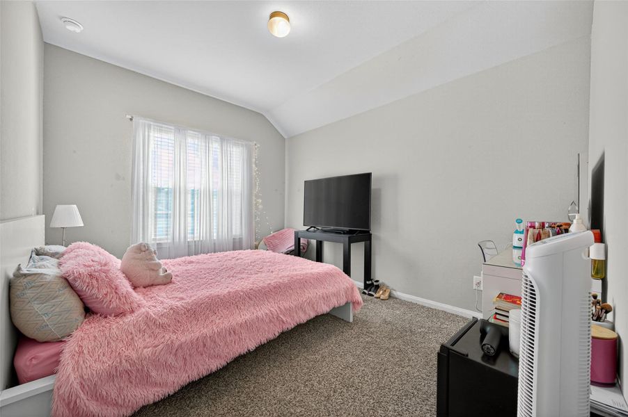 This cozy bedroom features soft carpeting, a large window with sheer curtains for natural light, and a plush bed with pink accents. The room includes a TV on a stand, a bedside lamp, and a small dresser, creating a comfortable and welcoming space.