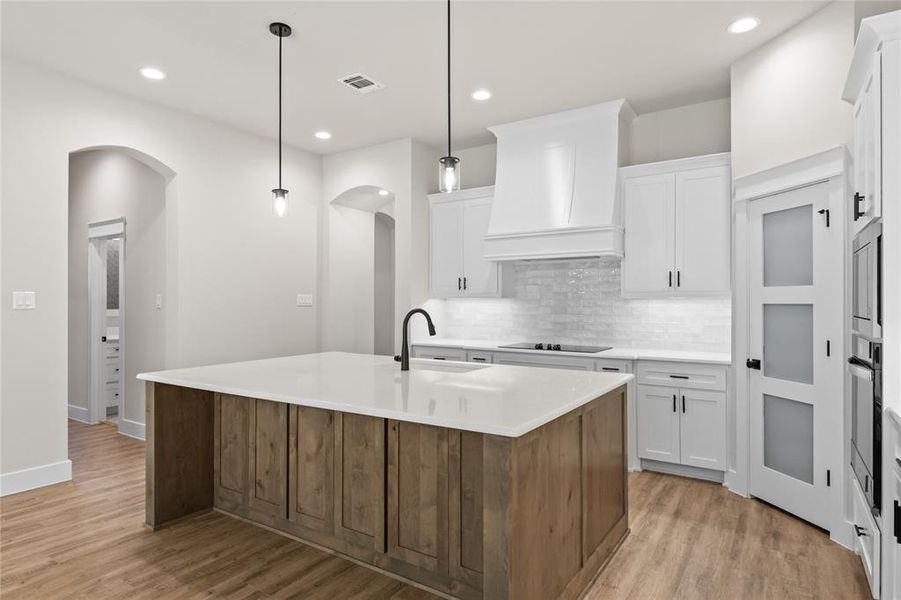 Two tone kitchen featuring a kitchen island with sink, arched walkways, two tone cabinets, light wood-type flooring, and hanging light fixtures