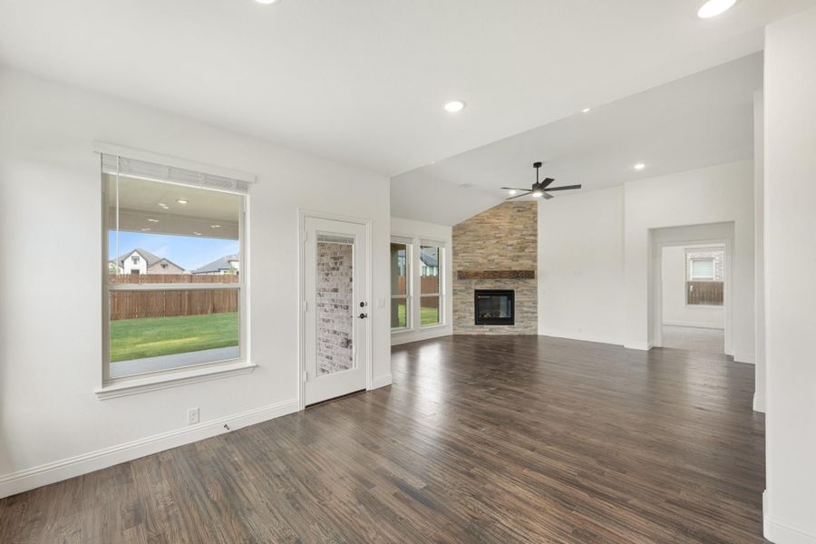 Open living room with dark hardwood floors, stone fireplace, ceiling fan, and backyard views through large windows.