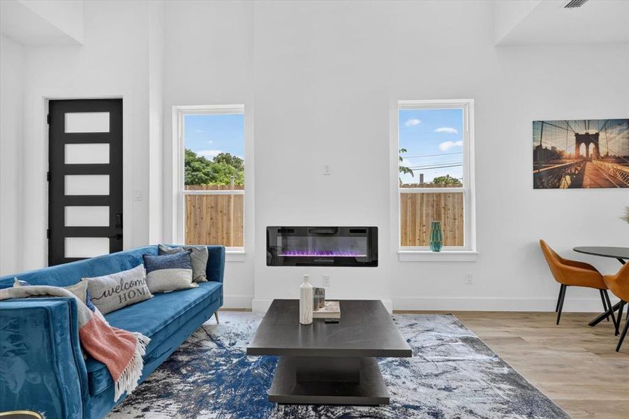 Open living area featuring vaulted ceilings, a contemporary wall-mounted electric fireplace, two large windows, a modern five-panel entry door, and wood-finish flooring