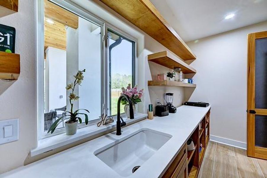 Bright and modern kitchen with large window, sleek countertops, and open wooden shelves. Ideal for cooking and entertaining, it offers ample natural light and a stylish, functional design.