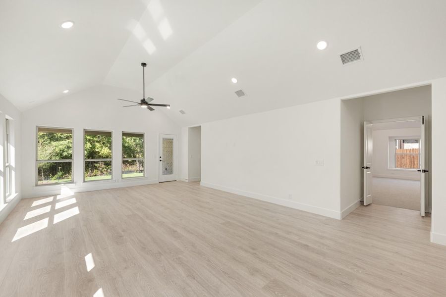 Open living room with vaulted ceiling, ceiling fan, light wood floors, and large windows overlooking backyard