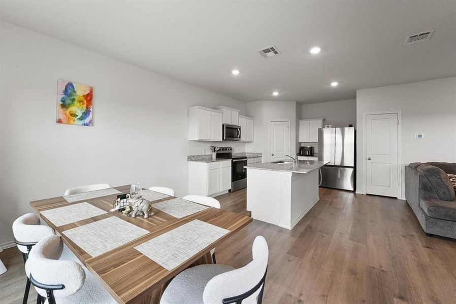 Open-concept living space featuring wood-finish flooring, recessed lighting, a kitchen island with sink, white shaker-style cabinetry, and stainless steel appliances
