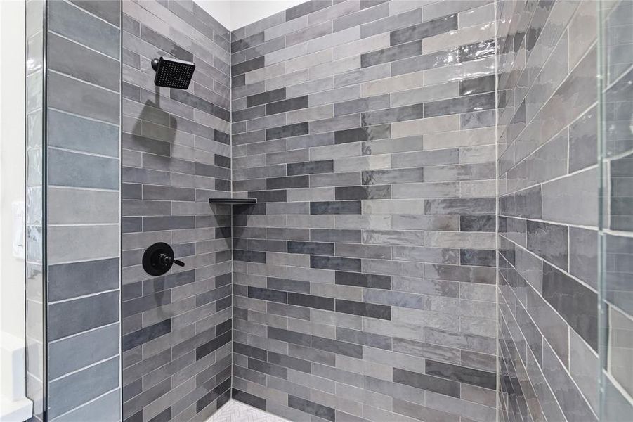Shower enclosure featuring variegated grey tile walls, a matte black rain shower head, coordinating matte black shower controls, a built-in shower shelf, and a glass partition