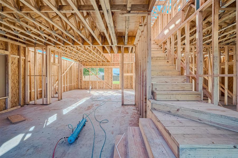 This image shows a home under construction with exposed wooden framing and an unfinished interior. Visible are a staircase and open spaces for future rooms. The structure receives ample natural light.