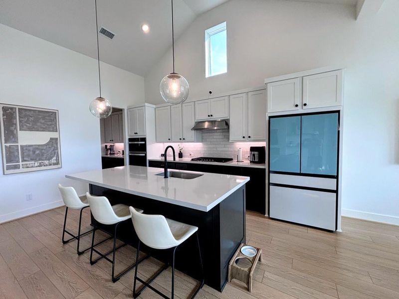 The kitchen features a modern design with high ceilings, white cabinetry, and a large central island with seating for three. It includes stainless steel appliances, stylish pendant lighting, and a light wood floor, creating an open and inviting space.