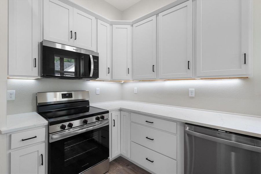 Kitchen featuring white shaker cabinetry, stainless steel appliances, quartz countertops, under-cabinet lighting, and wood-finish flooring