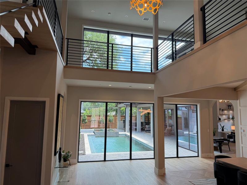 Double-height interior featuring light wood-finish flooring, a floating staircase with black metal railing, and extensive glass doors opening to a private pool area Double-height interior featuring light wood-finish flooring, a floating staircase with black metal railing, and extensive glass doors opening to a private pool area