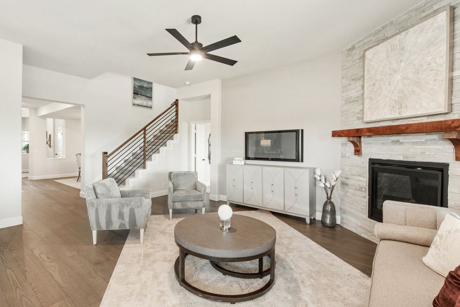 Living room with stone fireplace, hardwood floors, ceiling fan, and staircase with metal railing