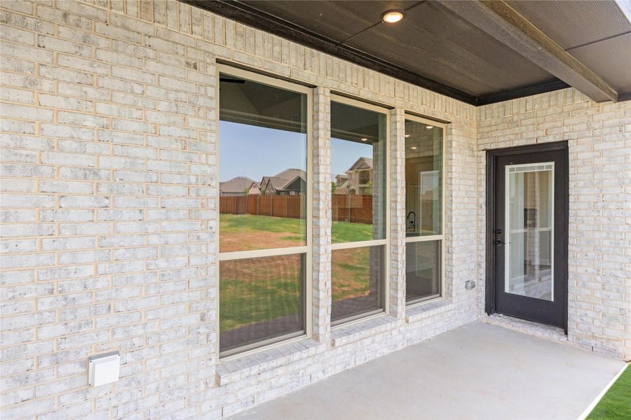 Covered back patio with large windows and glass door on light brick exterior wall overlooking fenced yard