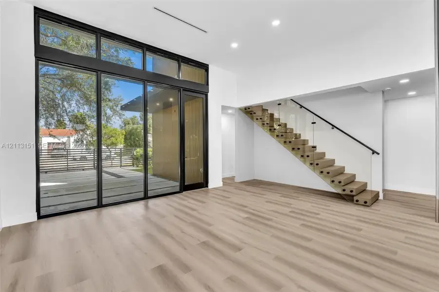 Floor-to-ceiling multi-panel glass doors, wood-finish flooring, and a floating staircase featuring glass railing panels and a black handrail Floor-to-ceiling multi-panel glass doors, wood-finish flooring, and a floating staircase featuring glass railing panels and a black handrail