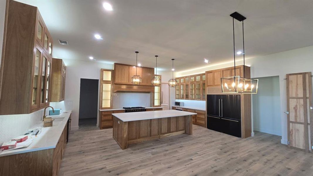 Kitchen with glass insert cabinets, brown cabinetry, a chandelier, high end black fridge, and decorative light fixtures