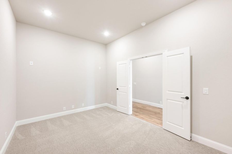 Empty bedroom with beige carpet, white walls, recessed lighting, and open door to hallway with hardwood floors Empty bedroom with beige carpet, white walls, recessed lighting, and open door to hallway with hardwood floors