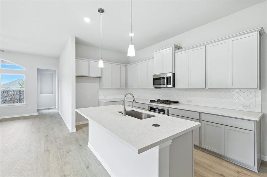 Kitchen with light wood finished floors, decorative light fixtures, a kitchen island with sink, backsplash, and stainless steel appliances Kitchen with light wood finished floors, decorative light fixtures, a kitchen island with sink, backsplash, and stainless steel appliances