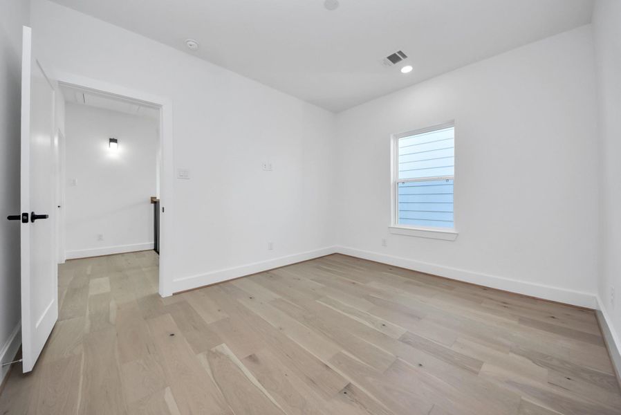 This room offers a minimalist design with light wood floors, white walls, and recessed lighting. A single window lets in natural light, while a white door connects to a well-lit hallway, offering versatility for a bedroom, office, or other use.