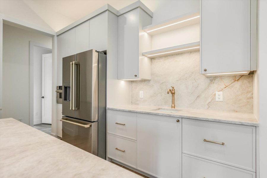 Modern kitchen featuring light cabinetry, integrated under-cabinet lighting, a stainless steel refrigerator, stone countertops, and a full-height stone backsplash with a brass faucet