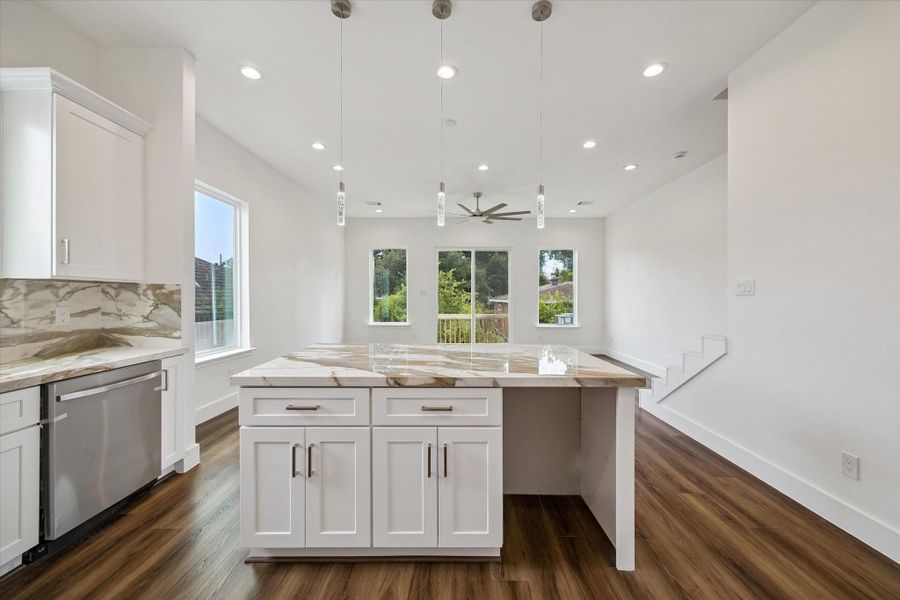 From the kitchen island, the open layout stretches toward the living area, showcasing the home’s easy flow and abundance of natural light. The broad stone-look island offers ample room for meal prep, serving, or casual seating, while pendant lights define the kitchen zone. Beyond, warm wood-look flooring, recessed lights, a modern ceiling fan, and multiple windows create a bright, cohesive space. A glass door opens to the outdoor area, adding another connection between inside and out. From the kitchen island, the open layout stretches toward the living area, showcasing the home’s easy flow and abundance of natural light. The broad stone-look island offers ample room for meal prep, serving, or casual seating, while pendant lights define the kitchen zone. Beyond, warm wood-look flooring, recessed lights, a modern ceiling fan, and multiple windows create a bright, cohesive space. A glass door opens to the outdoor area, adding another connection between inside and out.