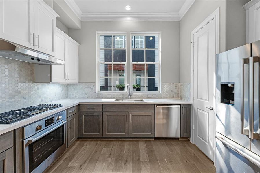 Recessed lighting throughout the space provides ample illumination, while large windows positioned above the stainless steel undermount sink flood the area with natural light. Gleaming wood floors add warmth and richness to the overall design, while elegant crown molding enhances the kitchen’s sophisticated aesthetic.