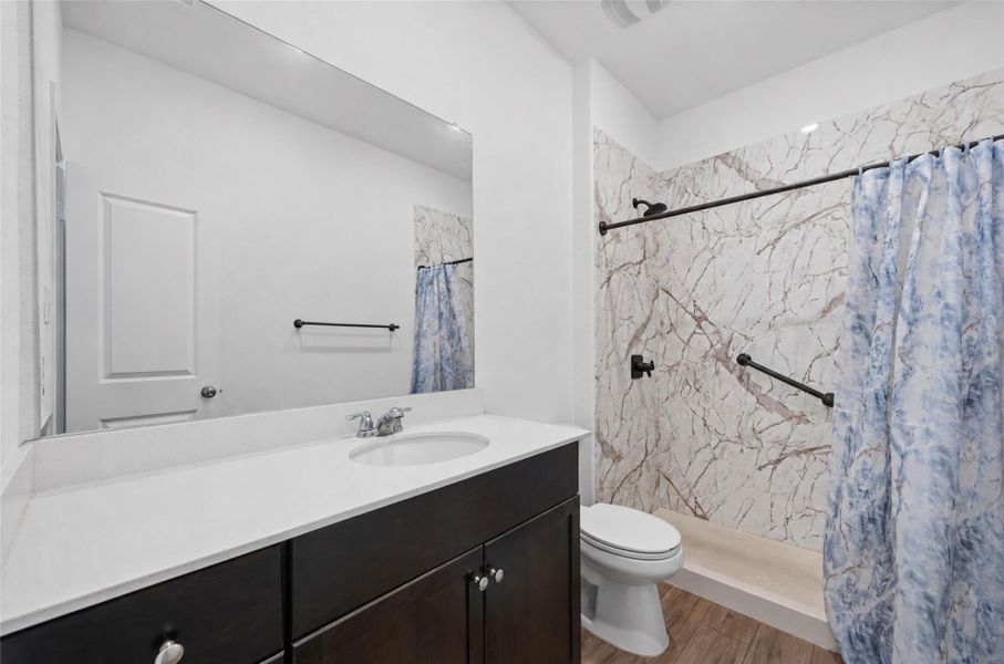 Bathroom featuring a dark wood-finish vanity with a white countertop and integrated sink, a large wall-mounted mirror, and a shower with patterned wall panels and matte black fixtures