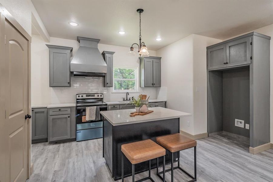Kitchen featuring gray cabinetry, stainless steel range with electric cooktop, backsplash, custom exhaust hood, and a kitchen bar