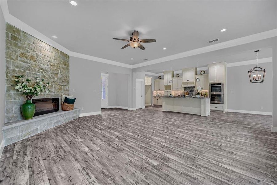 Unfurnished living room featuring wood-type flooring, a stone fireplace, ceiling fan with notable chandelier, and crown molding