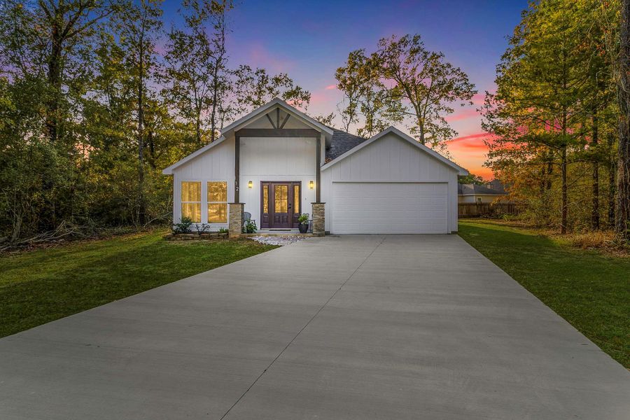 This photo showcases a charming single-story home with a modern design. It features a spacious front yard and a wide concrete driveway leading to a double garage. The exterior is highlighted by clean white siding, a welcoming front porch, and large windows. Surrounded by trees, the setting offers a serene and private atmosphere, enhanced by a vibrant sunset backdrop.
