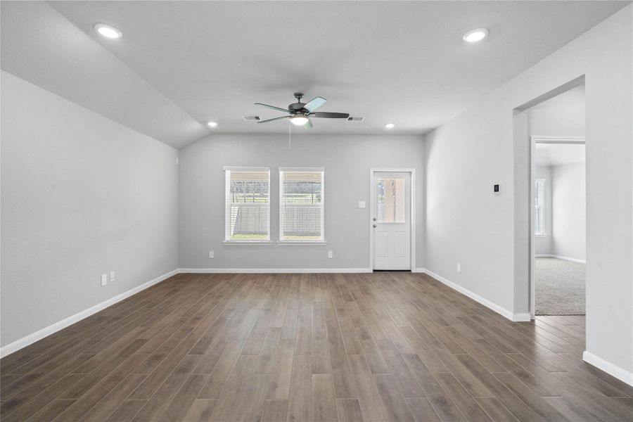 A bright and airy open-concept space featuring tile flooring, sloped ceilings with recessed lighting and ceiling fan, abundant natural light from large windows (fitted with blinds), and an access door to the back covered patio.