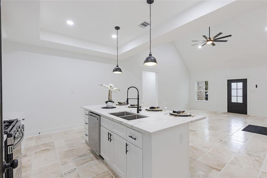 This is a spacious, modern kitchen featuring high vaulted ceilings with recessed lighting, sleek pendant lights, a large central island with a sink, and stainless steel appliances. The room is finished with tiled flooring and has plenty of natural light from windows and a glass door leading outside.