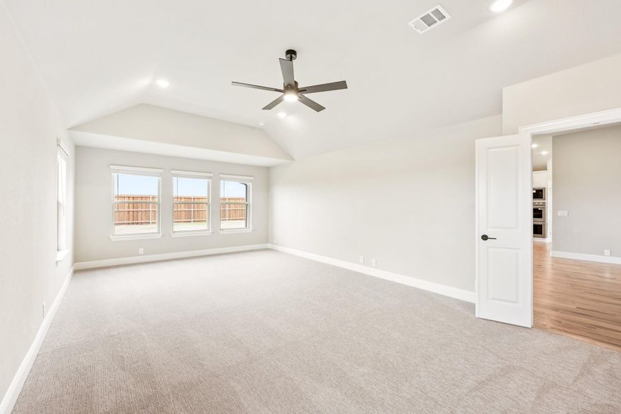 Empty living room with vaulted ceiling, ceiling fan, carpet flooring, and triple windows overlooking a fenced backyard. Empty living room with vaulted ceiling, ceiling fan, carpet flooring, and triple windows overlooking a fenced backyard.